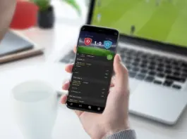 En person håller i en mobil med en bettingapp, i bakgrunden syns en laptop med en fotbollsmatch