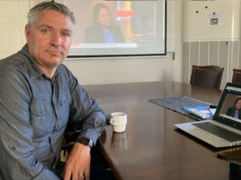 Man vid konferensbord med laptop, kaffekopp och projektorbild i bakgrunden.