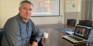 Man vid konferensbord med laptop, kaffekopp och projektorbild i bakgrunden.