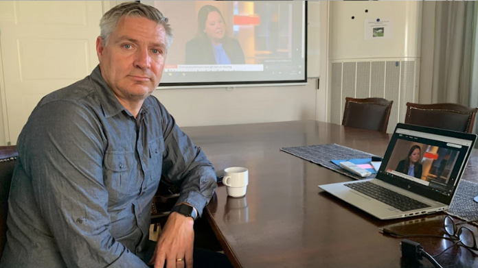 Man vid konferensbord med laptop, kaffekopp och projektorbild i bakgrunden.