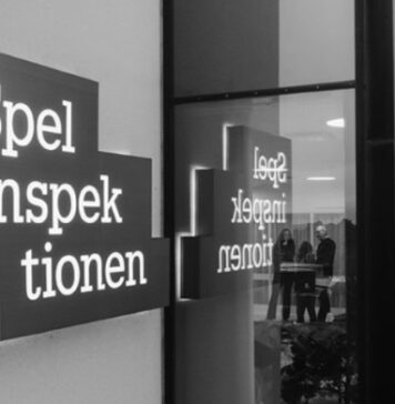 Skylt med texten Spelinspektionen utanför en byggnad, speglas i fönsterglas med personer synliga inomhus.