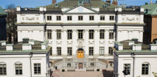 Vit stenbyggnad med grönt tak och stora fönster, pampig entré med trappa och gula dörrar.