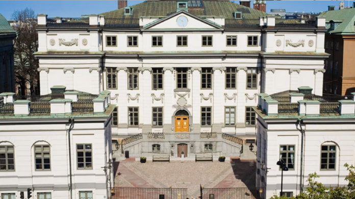 Vit stenbyggnad med grönt tak och stora fönster, pampig entré med trappa och gula dörrar.