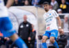 En fotbollsspelare i vit och blå matchdräkt under en match, med publiken suddig i bakgrunden.