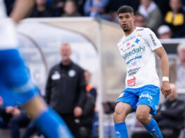 En fotbollsspelare i vit och blå matchdräkt under en match, med publiken suddig i bakgrunden.
