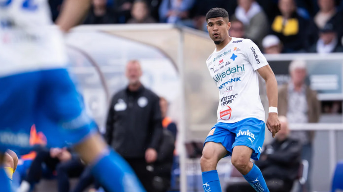 En fotbollsspelare i vit och blå matchdräkt under en match, med publiken suddig i bakgrunden.